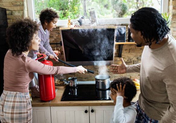 African American family using fire extinguisher while spraying smoke from a saucepan in the kitchen. Focus is on shocked girl.
